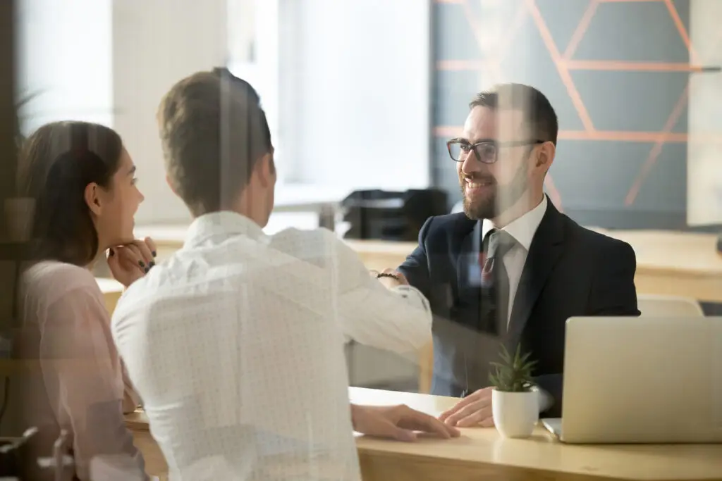 Smiling male realtor or broker shaking hand of excited buyers couple, negotiating about first house purchase or taking loan, insurance agent welcoming clients with handshake at consultation meeting